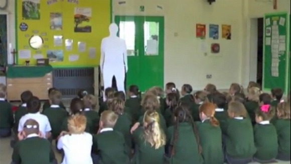 Children in assembly gaze up at the cardboard clergyman.
