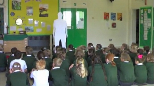 Children in assembly gaze up at the cardboard clergyman.
