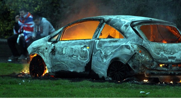 A man wrapped in a Union flag sits beside a burnt out car in Belfast during the flag protests.