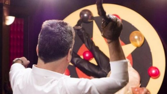 Simon Cowell in the foreground with a whip, while David Walliams is tied to a rotating wheel