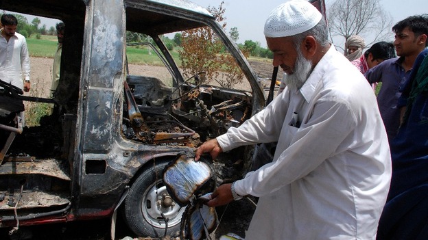 A man displays a burnt book to the media near a burnt-out school bus on the outskirts of Gujrat