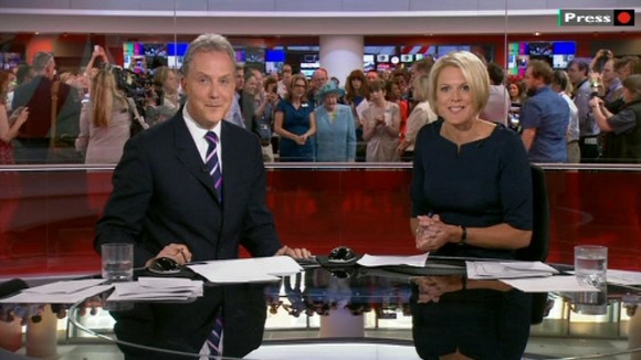 The BBC News presenters seem unaware that the Queen is behind them during their broadcast.