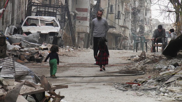 Civilians stand along a street lined with rubble in the besieged area of Homs